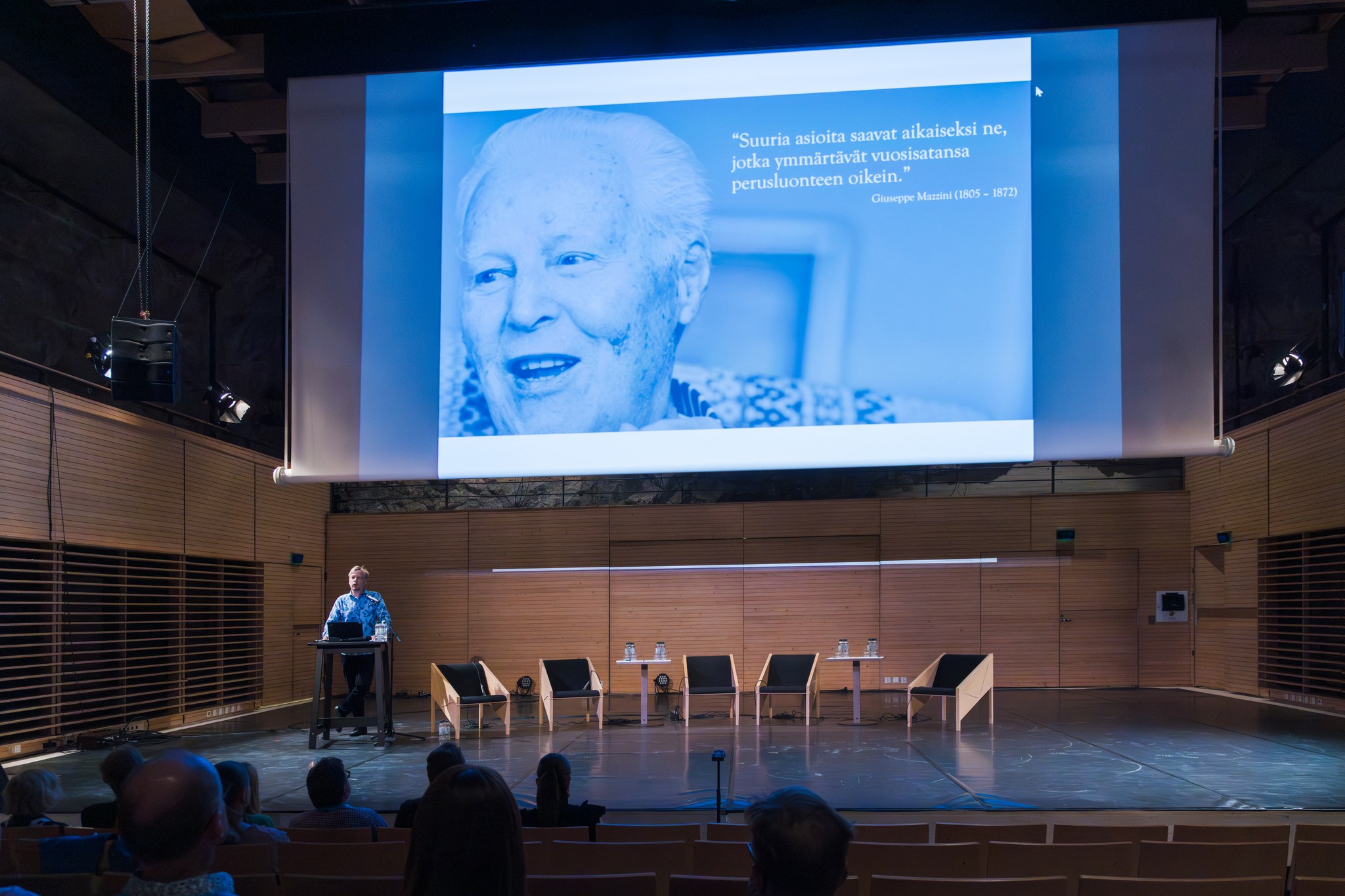 kuvassa edesmenneen festivaalien presidentin suuri kuva Kaustinen-salin screenillä Antti Huntuksen pitäessä puhetta kulttuurista elinvoimaa -seminaarissa.