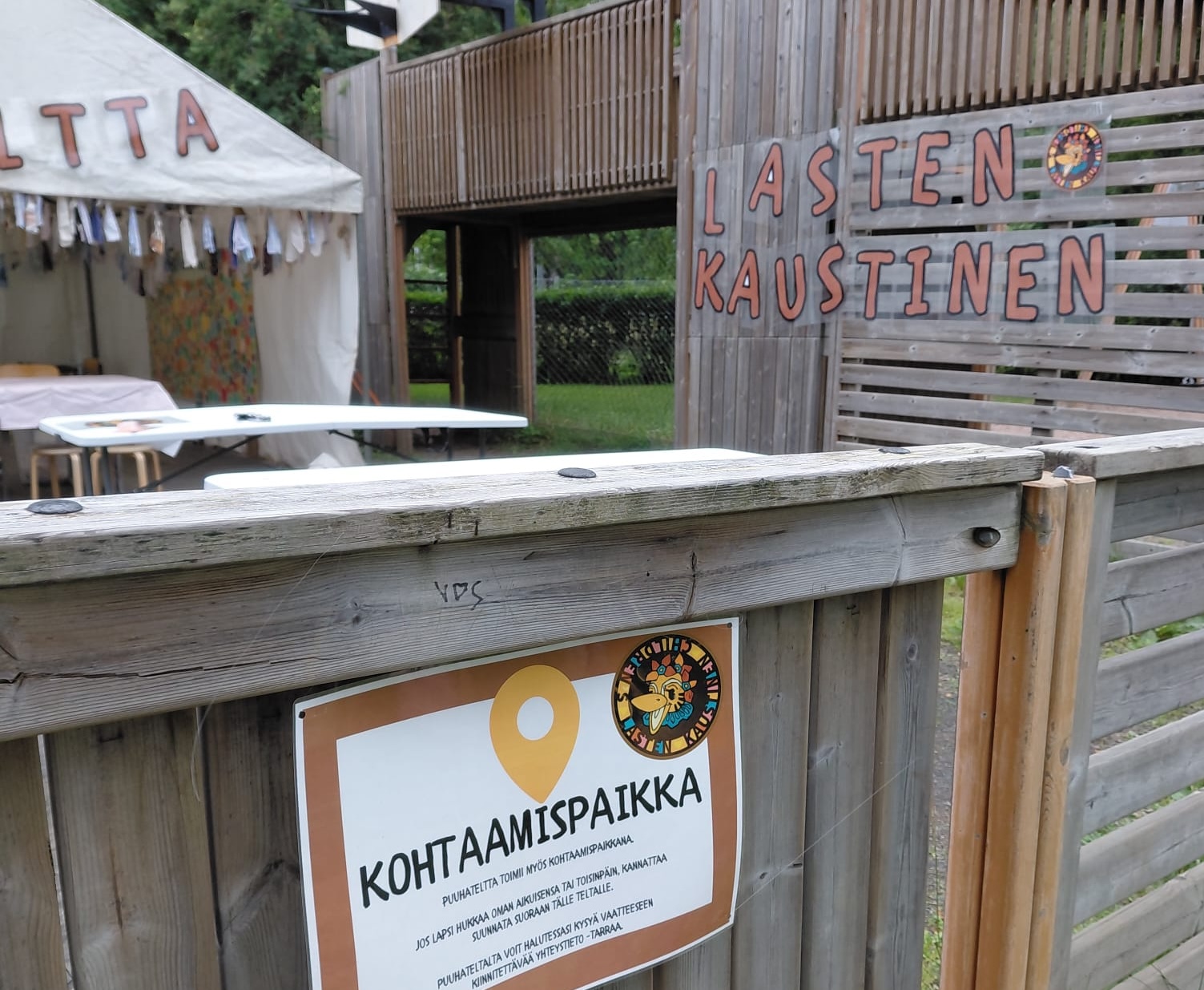 Kohtaamispistekyltti aidassa, taustalla näkyy lasten kaustinen -teksti.